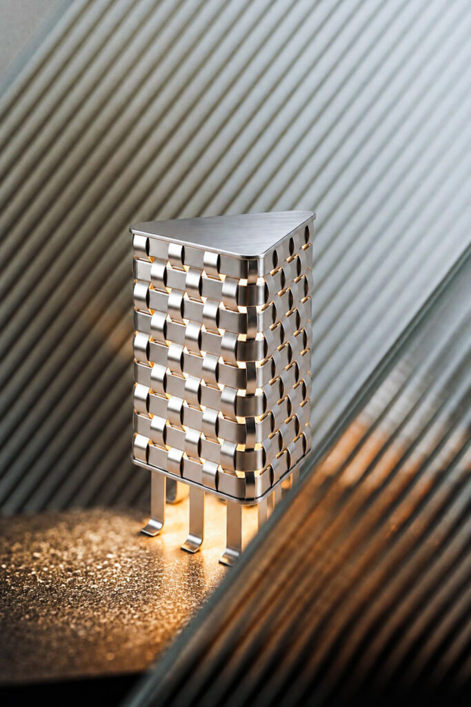 OT lamp in an interior – woven stainless-steel table lamp glowing beside ribbed glass